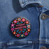 Round black pinback button with vibrant floral embroidery design surrounding bold pink and red text reading "Dissent is Patriotic."